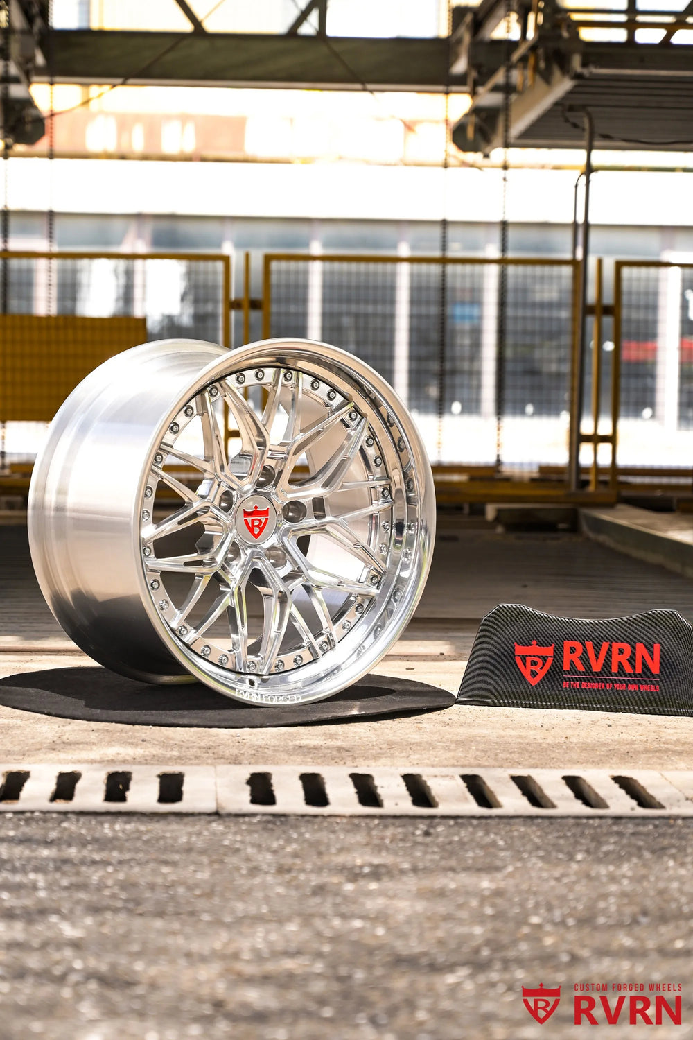 1970 Chevy Nova RV-T081 2-piece forged Y-Spoke Deep Dish wheel with Polished Spoke, Polished Flange, Polished Barrel, and RVRN Red Screws, presented in a wide workshop composition with the wheel upright on a black mat, deep polished barrel, red center emblem, RVRN sign, and industrial background visible.