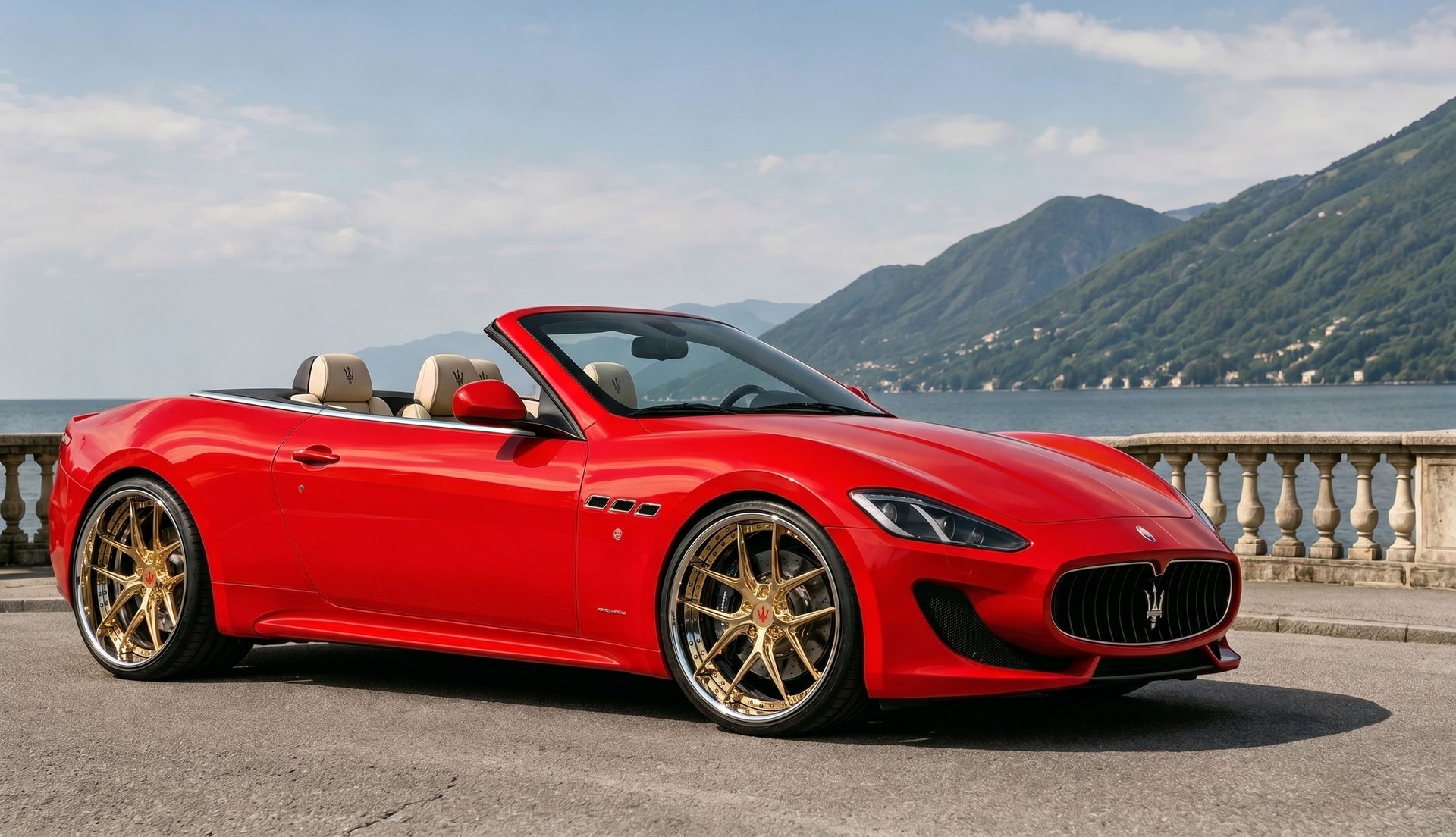A red Maserati GranTurismo convertible sports car parked outdoors on a paved area with a scenic lake and mountains in the background. The car is shown from a three-quarter front angle with its top down, featuring gold multi-spoke wheels and beige leather seats.