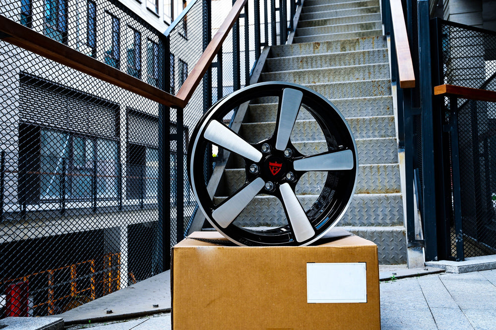 RVRN RV-MP223 forged monoblock wheel for a 2011 Chevy Camaro LT photographed on a shipping box beside an exterior staircase, brushed spokes and gloss black barrel.
