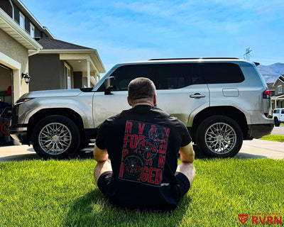 2026 Lexus GX 550 on RVRN RV-MG122 custom forged wheels in Gloss Gunmetal, owner in RVRN FORGED shirt sitting on the lawn beside the SUV.