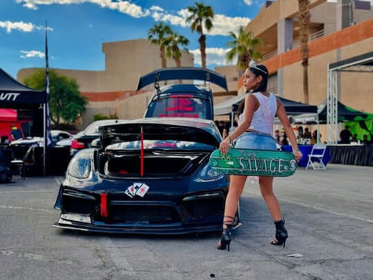 Woman posing with skateboard next to a modified Porsche Cayman, showcasing custom wheels at a car event.