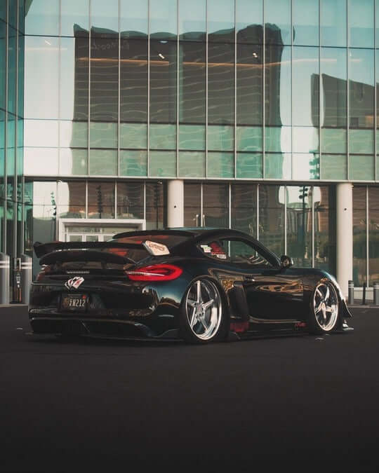 Custom Porsche Cayman with forged wheels and 5 spoke deep lip rims, showcased against a modern glass building backdrop.
