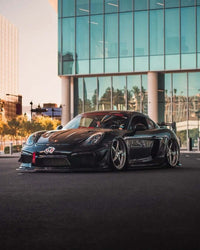 Custom Porsche Cayman with forged wheels and 5 spoke deep lip rims parked outside a modern building under clear skies.
