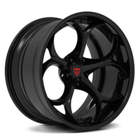 A glossy black multi-spoke alloy wheel rim with a red shield logo badge in the center, shown on a clean white background.