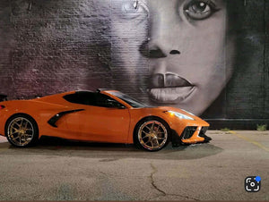 Side profile of an orange 2024 Corvette C8 Stingray on RVRN RV-DR08 deep dish gold chrome wheels parked before a night mural
