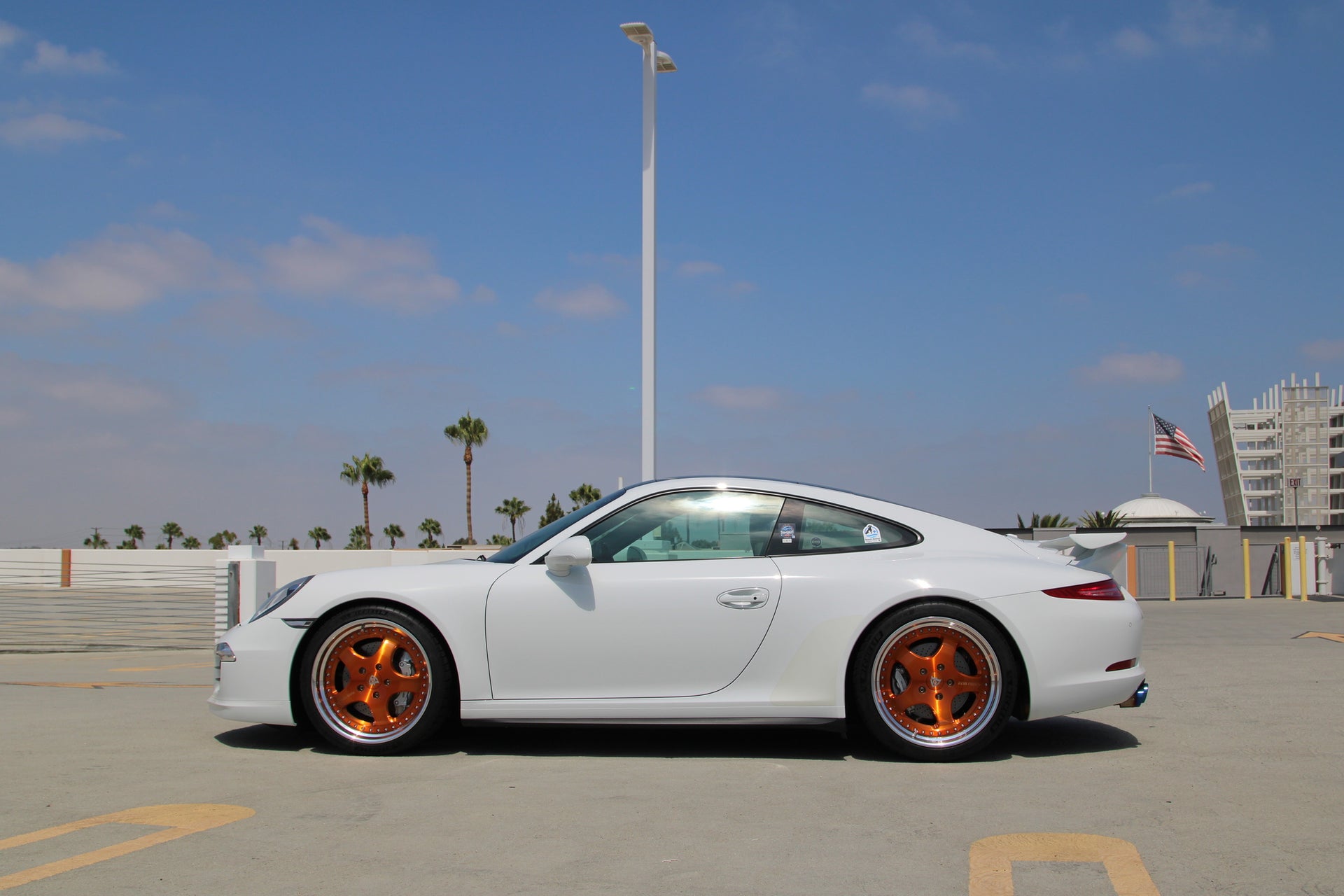 A white Porsche 911 coupe with distinctive orange multi-spoke wheels, parked outdoors on a paved surface under a blue sky with palm trees and a building in the background.