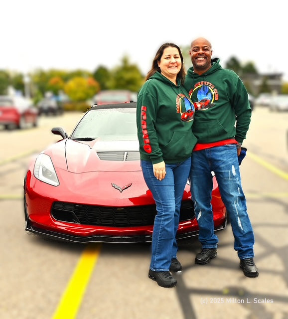 Two people in matching green sweatshirts standing next to a red corvette sports car on a road.