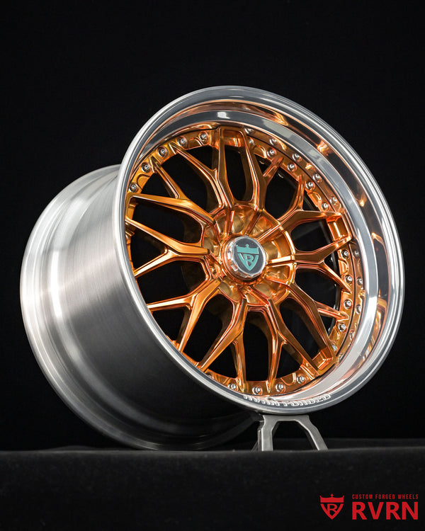 A multi-spoke alloy wheel rim with a polished silver outer barrel and a highly reflective copper-orange inner face, displayed on a stand against a black background. The center cap features a small green shield logo, and an RVRN watermark is visible in the bottom right corner.