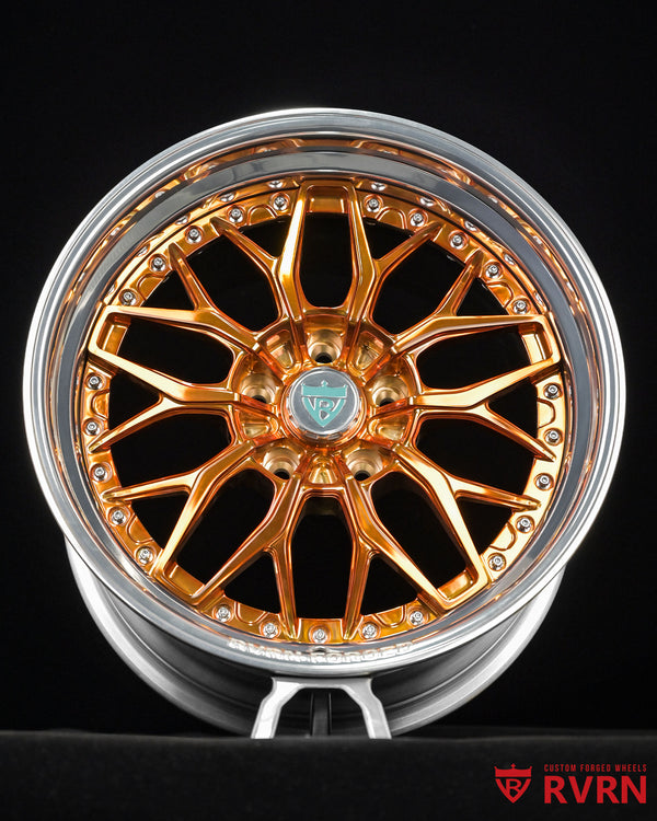 A close-up studio shot of a multi-spoke alloy wheel rim with a polished gold finish and a silver outer lip, shown against a black background. The center cap features a silver and green emblem, and the outer barrel has a row of small clear stones.